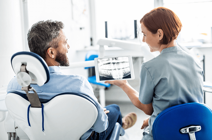 A dentist sits next to a patient in a dental chair and shows him X-rays of his teeth.