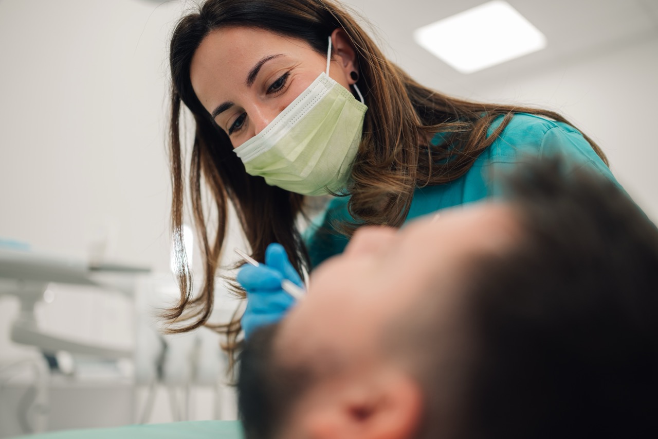 Female dentist wearing a surgical mask and blue gloves, carefully examining the teeth of a male patient reclining in a dental chair at a modern dental clinic