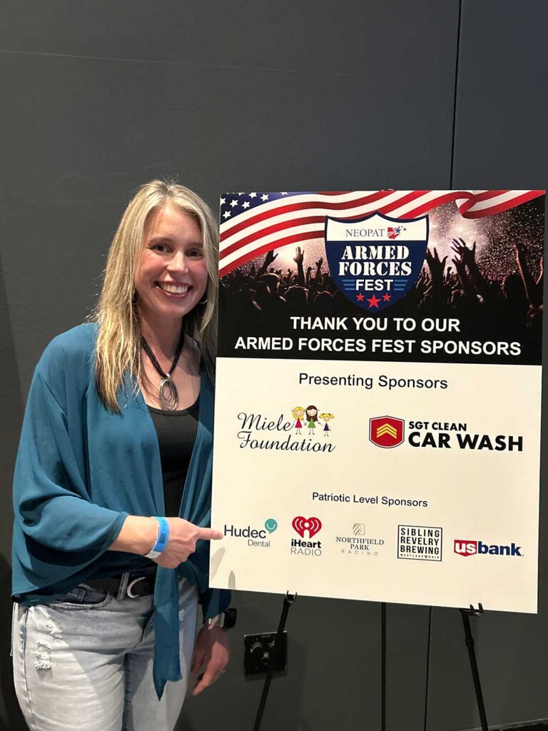 woman smiling with sign that says thank you to our armed forces fest sponsors with the hudec logo on it
