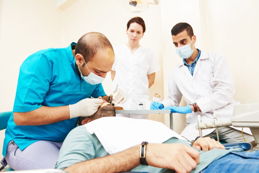 A dentist and student observe a dental assistant cleaning a patient's teeth