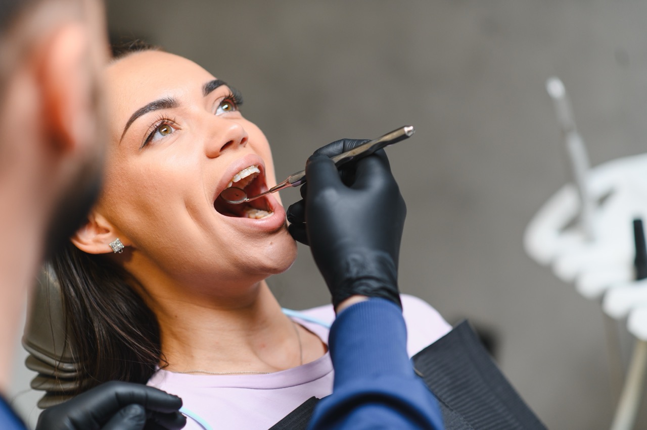 Dentist in black gloves examining a female patient's open mouth with a dental mirror during a routine checkup
